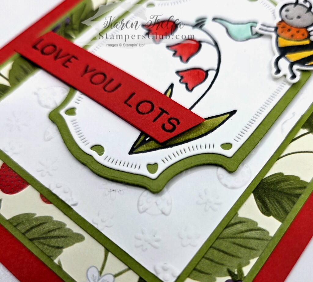Angled view of a Sweet Bugs Stampin' Up card featuring a bee watering red bell flowers with a Love You Lots sentiment strip on a red card base with embossed texture and berry print designer paper